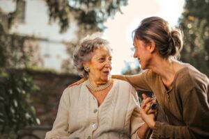 A young woman with her hair in a bun smiles warmly while holding hands with an elderly woman wearing a pearl necklace and a cream-colored cardigan. They are outdoors, surrounded by greenery, sharing a heartfelt moment.