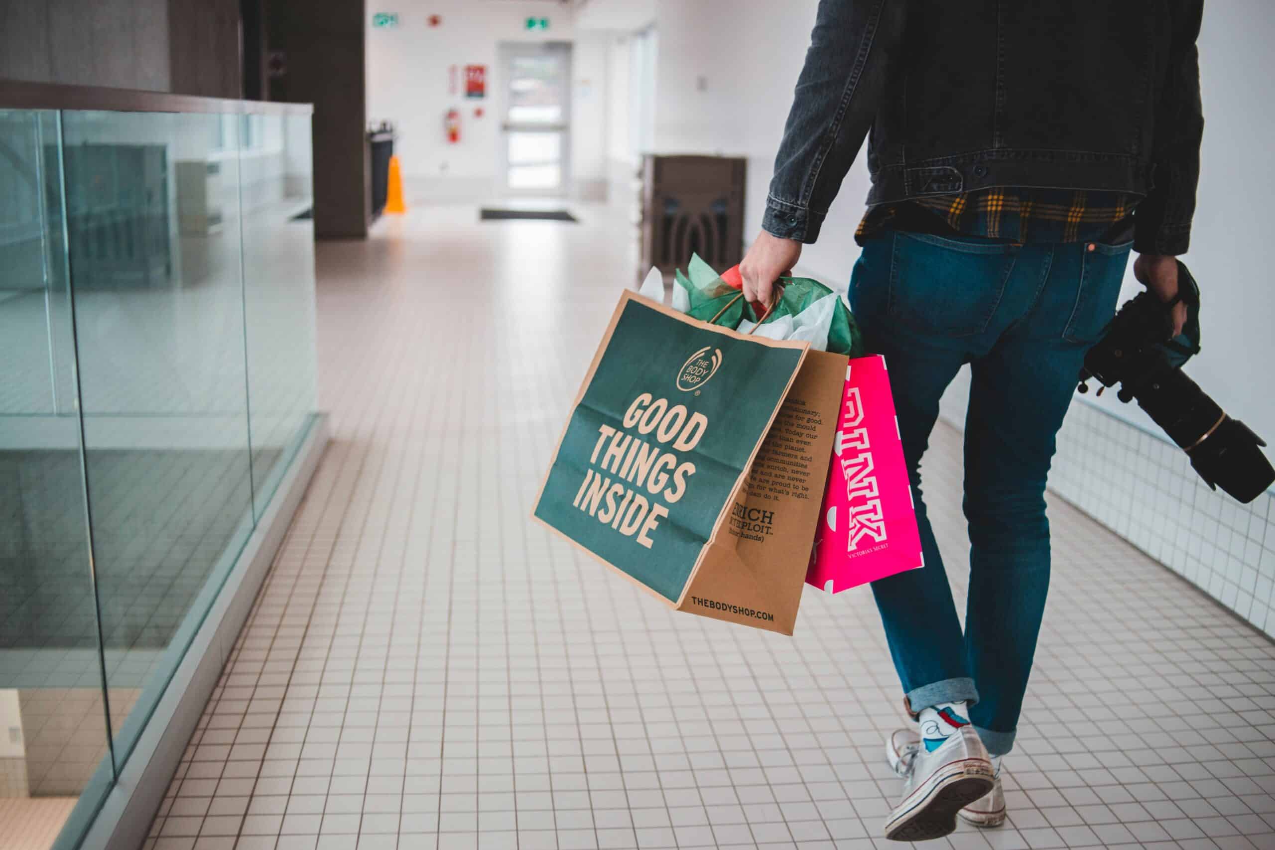 A person walking down a tiled indoor hallway holding shopping bags from The Body Shop and PINK in one hand, and a professional camera in the other. The focus is on the lower half of the person wearing jeans, sneakers, and a plaid shirt under a black jacket. The green bag reads “Good Things Inside.” The setting suggests a mall or shopping center.